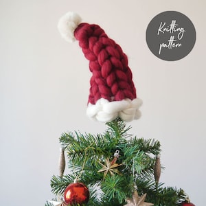 May include: A red and white knitted Santa hat tree topper. The hat is made with chunky yarn and has a white pom pom on top.