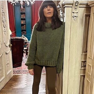 May include: A woman wearing an olive green, chunky knit sweater with a high neckline and matching leggings. She is also wearing brown suede cowboy boots. The outfit is in a doorway with a wooden floor.