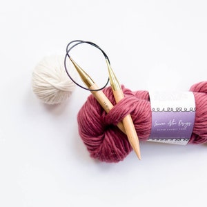 Puede incluir: Dos agujas de tejer de madera con puntas doradas y un lazo de cord&oacute;n negro est&aacute;n envueltas alrededor de una bola de hilo rosa oscuro. El hilo est&aacute; etiquetado como "Lauren Aston Designs Super Chunky Yarn".