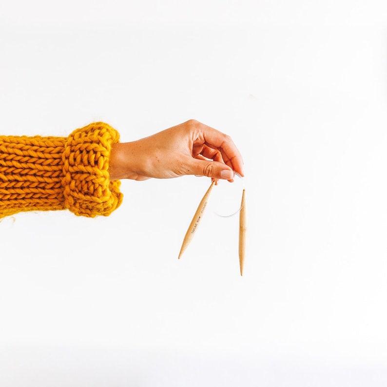 May include: A hand holding two wooden knitting needles with a strand of yarn attached. The hand is wearing a yellow knitted sweater.