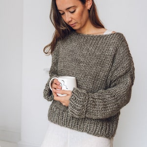 May include: A woman wearing a chunky knit, oversized, grey sweater with a white mug in her hands.