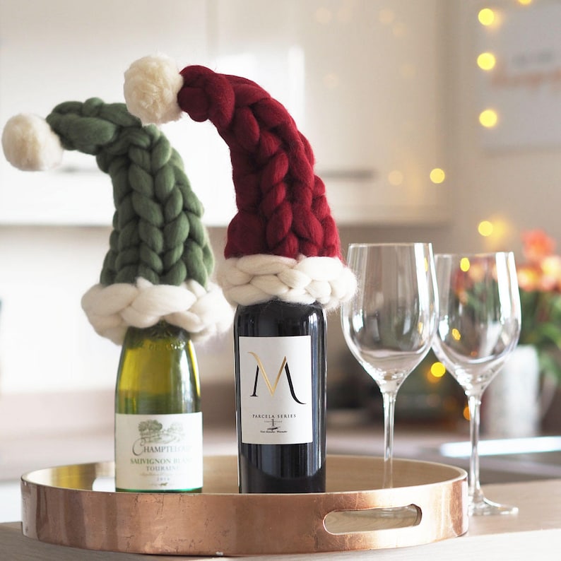 May include: Two wine bottles with knitted Santa hats. The bottle on the left is green and has a white label with the text "Champs Vineyards Sauvignon Blanc 2019". The bottle on the right is red and has a white label with the text "Parcela Series M".