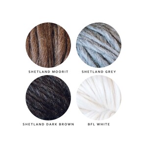 K&ouml;nnte beinhalten: Vier Kn&auml;uel Garn in verschiedenen Farben: Braun, Grau, Schwarz und Wei&szlig;. Das Garn ist mit "Shetland Moorit", "Shetland Grey", "Shetland Dark Brown" und "BFL White" beschriftet.