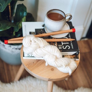 K&ouml;nnte beinhalten: Ein Kn&auml;uel wei&szlig;er Wolle mit zwei Holzstricknadeln mit roten Spitzen. Die Wolle liegt auf einem Holzhocker mit einem Buch mit dem Titel "Studio" darauf. Eine Tasse Kaffee steht auf dem Buch.