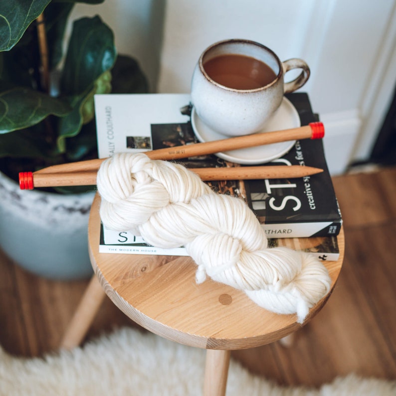K&ouml;nnte beinhalten: Ein Kn&auml;uel wei&szlig;er Wolle mit zwei Holzstricknadeln mit roten Spitzen. Die Wolle liegt auf einem Holzhocker mit einem Buch mit dem Titel "Studio" darauf. Eine Tasse Kaffee steht auf dem Buch.