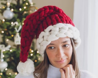 May include: A chunky knit Santa hat featuring a deep red main section with a thick, white braided brim and a large white pom-pom at the tip. The hat is constructed from thick, soft yarn with a prominent, textured weave throughout the entire design.