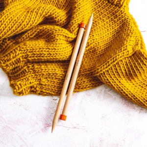 Puede incluir: Dos agujas de tejer de madera con puntas naranjas, sobre una superficie blanca con un su&eacute;ter de punto amarillo en el fondo.
