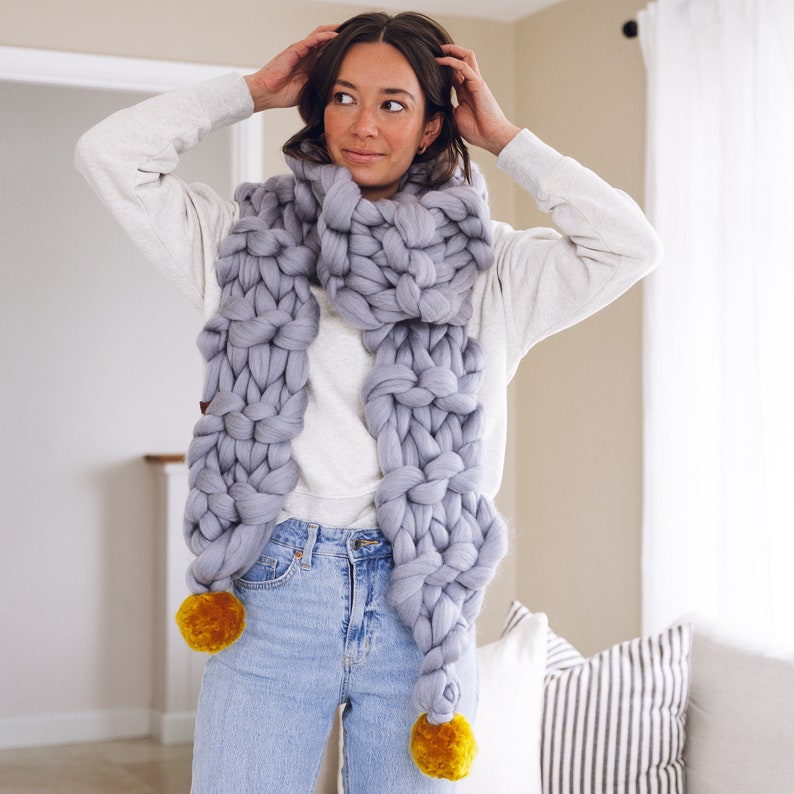 May include: A chunky knit scarf in a light gray color with two yellow pom-poms at the ends. The scarf is worn by a person with a white sweater and blue jeans.