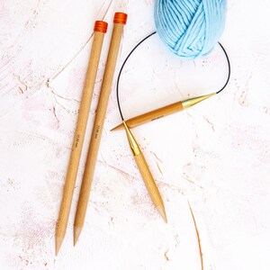 Puede incluir: Dos agujas de tejer de madera con puntas naranjas y una aguja de tejer circular con punta dorada. Las agujas est&aacute;n sobre una superficie blanca texturizada con un ovillo de lana azul claro.