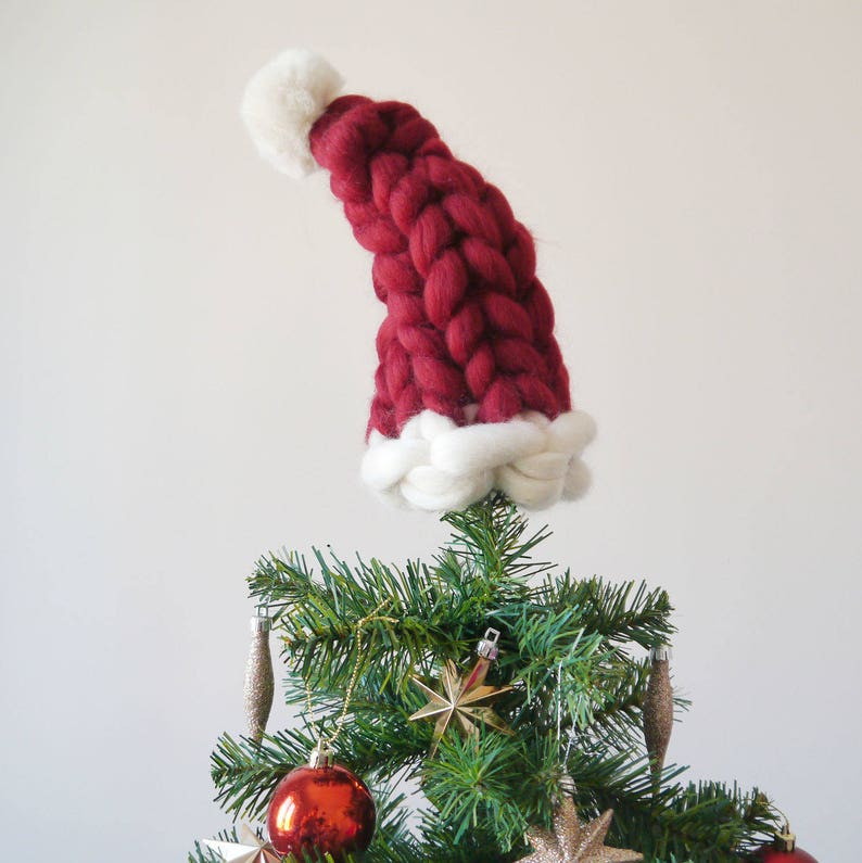 May include: A red knitted Santa hat with a white pom-pom and white trim sits atop a green Christmas tree.
