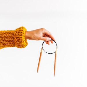 May include: A pair of wooden knitting needles with gold tips and a black cord connecting them. The needles are held in a hand wearing a yellow knitted sweater.