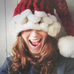 May include: A woman wearing a red and white knitted Santa hat with a white pom-pom. She is smiling and her eyes are closed. The hat is pulled down over her eyes.