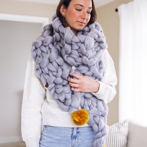 May include: A woman wearing a chunky knit scarf with a large yellow pom-pom. The scarf is a light gray color and is made of thick yarn.