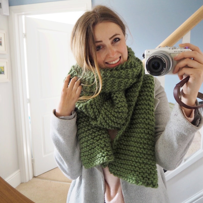 May include: A woman wearing a gray coat and a green knitted scarf. She is holding a white camera in her right hand.