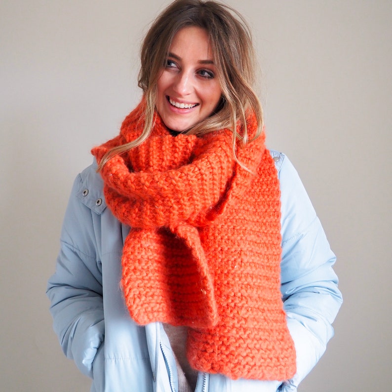 May include: A woman wearing a light blue jacket with a bright orange knitted scarf. The scarf is thick and chunky, and the woman is smiling.
