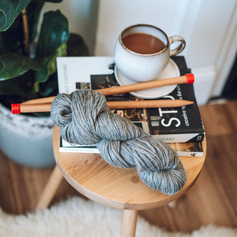 K&ouml;nnte beinhalten: Ein graues Garnkn&auml;uel mit zwei Stricknadeln aus Holz und einer roten Spitze an jeder Nadel. Das Garn liegt auf einem Holzhocker mit einem Buch mit dem Titel "The Modern House" und einer Tasse Kaffee.