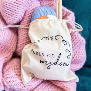 May include: A white cotton drawstring bag with the text "PURLS OF wisdom" printed in black. The bag is filled with a ball of light blue yarn and a wooden knitting needle.