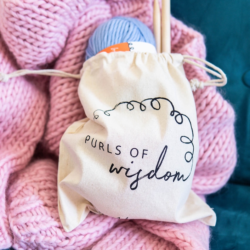 May include: A white cotton drawstring bag with the text "PURLS OF wisdom" printed in black. The bag is filled with a ball of light blue yarn and a wooden knitting needle.