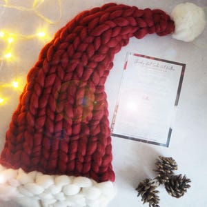 May include: A large, red knitted Santa hat with a white pom-pom on top. The hat is made with chunky yarn and has a white fur trim around the bottom.