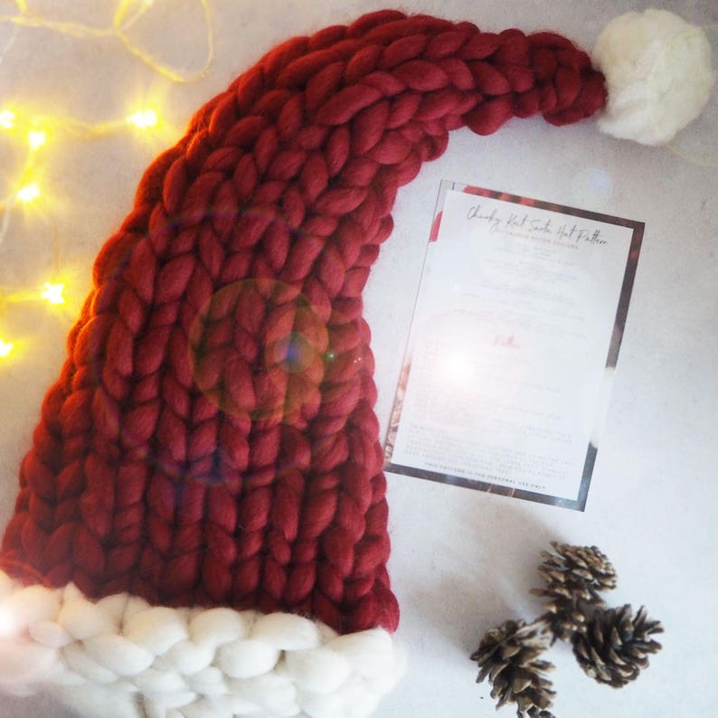 May include: A large, red knitted Santa hat with a white pom-pom on top. The hat is made with chunky yarn and has a white fur trim around the bottom.