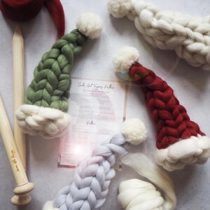 May include: Three knitted Santa hats in green, red, and gray with white pom-poms. The hats are lying on a white surface with a piece of paper with the text "Santa Hat Tipping Pattern" on it.