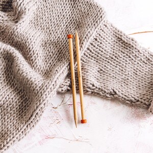 May include: Two wooden knitting needles with orange tips, lying on a white surface with a beige knitted fabric in the background. The needles are labeled "10.00 mm" and "12.00 mm".