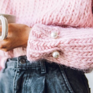 Könnte beinhalten: Ein rosa gestrickter Pullover mit Perlenknöpfen an den Ärmeln. Der Pullover ist weich und flauschig, und die Perlenknöpfe verleihen ihm einen Hauch von Eleganz.