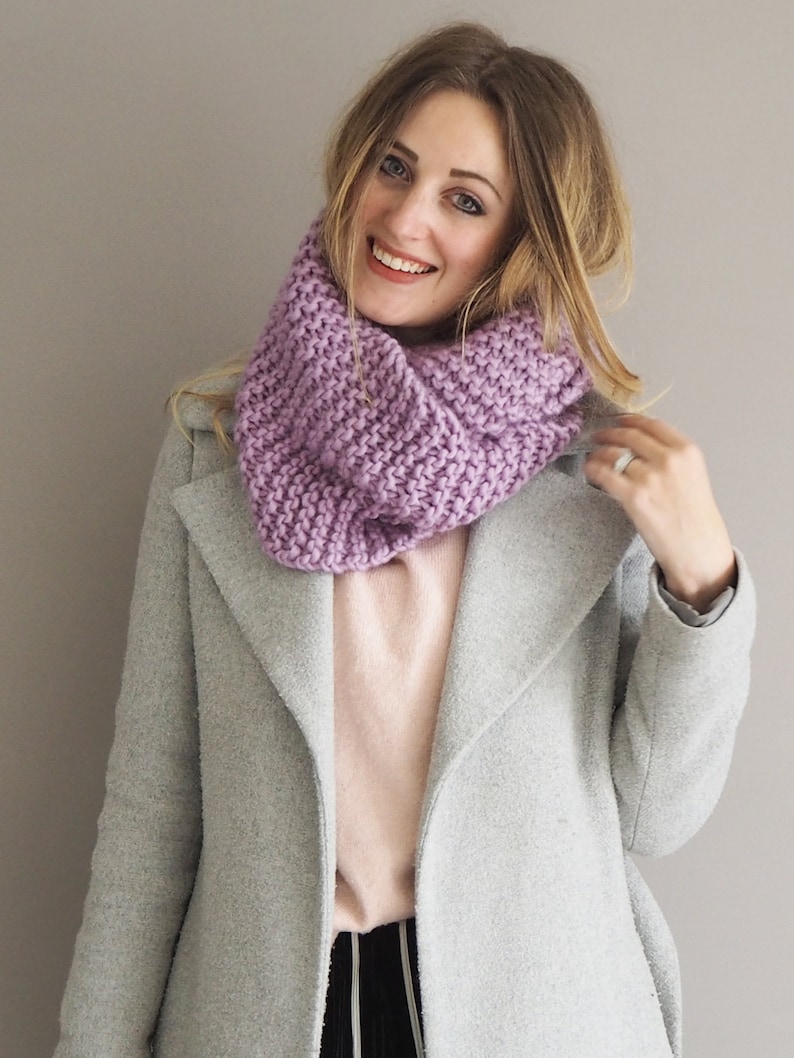 May include: A woman wearing a light gray coat with a purple infinity scarf. The scarf is knitted and has a textured pattern.