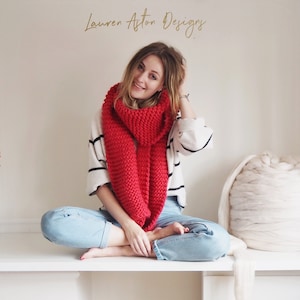 May include: A woman wearing a red chunky knit scarf, a white and black striped sweater, and blue jeans. The scarf is draped around her neck and is very thick. The woman is sitting on a white surface with a large white yarn ball behind her.