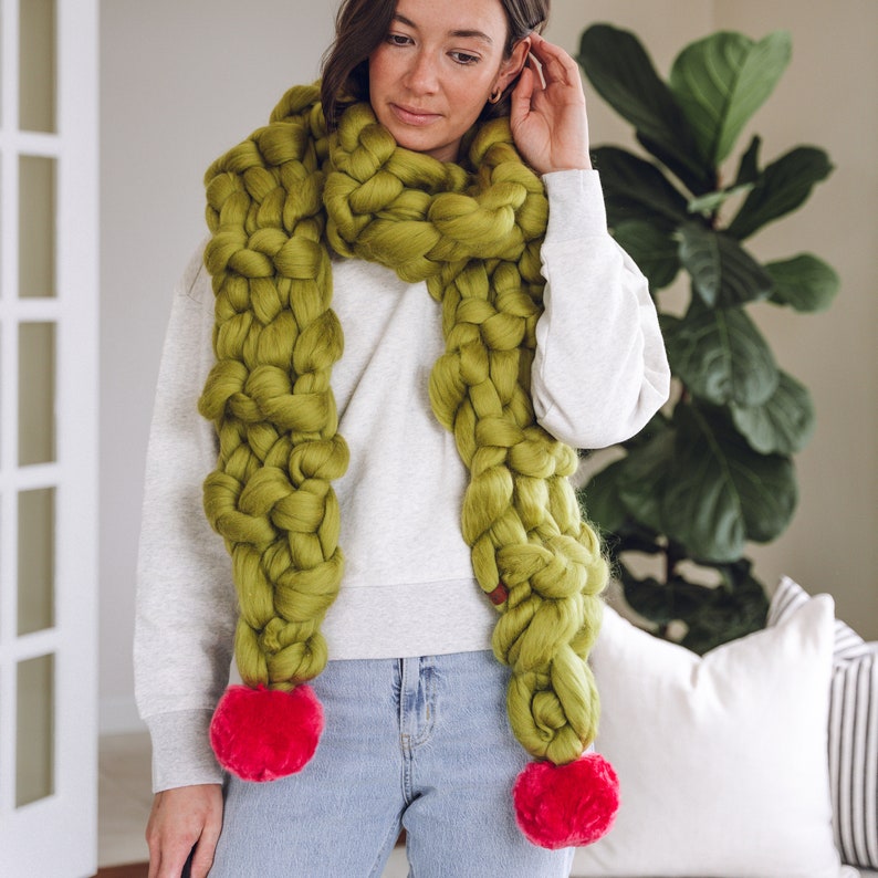 May include: A chunky knit scarf in a bright green color with two large pink pom-poms on the ends. The scarf is worn around the neck of a person wearing a white sweater and blue jeans.
