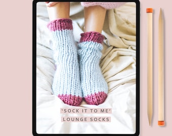 May include: A digital tablet displaying a knitting pattern for 'Sock It To Me' lounge socks. The image shows a pair of blue and pink knitted socks on a white bedspread. The text 'Lauren Acton Designs' is at the bottom of the screen.