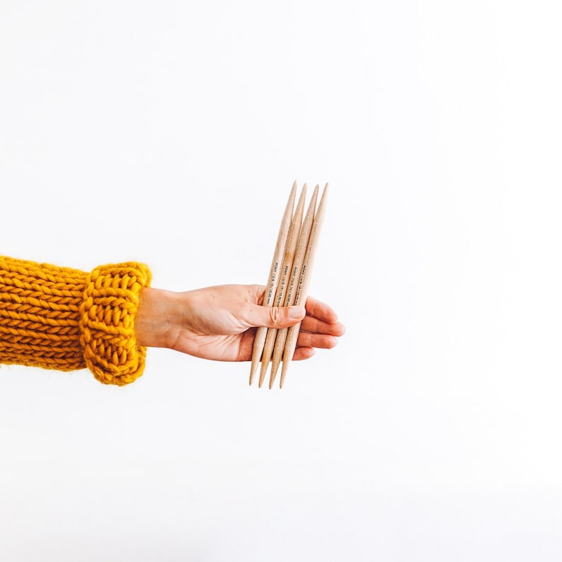 May include: A hand holding a set of five wooden knitting needles. The needles are all the same size and have a smooth finish.