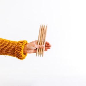 以下が含まれることがあります： 手には、先端が尖った5本の木製の編み針があります。 針は薄茶色で、サイズを示す黒い文字（12.00 mm）が書かれています。 背景は白で、黄色のニットスリーブが見えます。