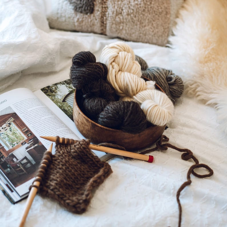 K&ouml;nnte beinhalten: Eine Holzschale gef&uuml;llt mit verschiedenen Braunt&ouml;nen und cremefarbenen Garnen, darunter eine Kn&auml;uel braunes Garn auf einer Stricknadel. Die Schale steht auf einem wei&szlig;en Bettlaken mit einem Magazin und einem gestrickten Stoffst&uuml;ck.