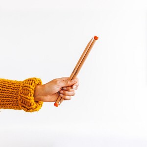 May include: A hand holding two wooden knitting needles with orange tips. The needles are crossed and the hand is wearing a yellow knitted sweater.