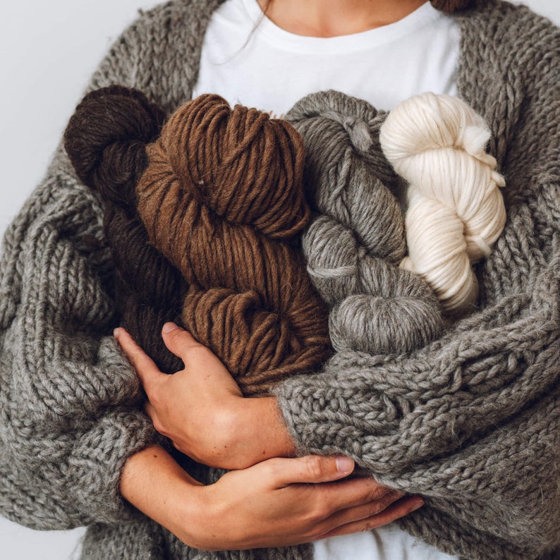 K&ouml;nnte beinhalten: Eine Person tr&auml;gt einen grauen Strickcardigan und h&auml;lt verschiedene Garnkn&auml;uel in verschiedenen Farben und Texturen, darunter Braun, Grau und Wei&szlig;.