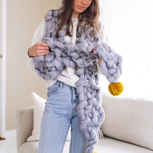 May include: A woman wearing a light gray chunky knit scarf with a yellow pom-pom. The scarf is draped around her neck and shoulders. She is wearing blue jeans and a white sweater.