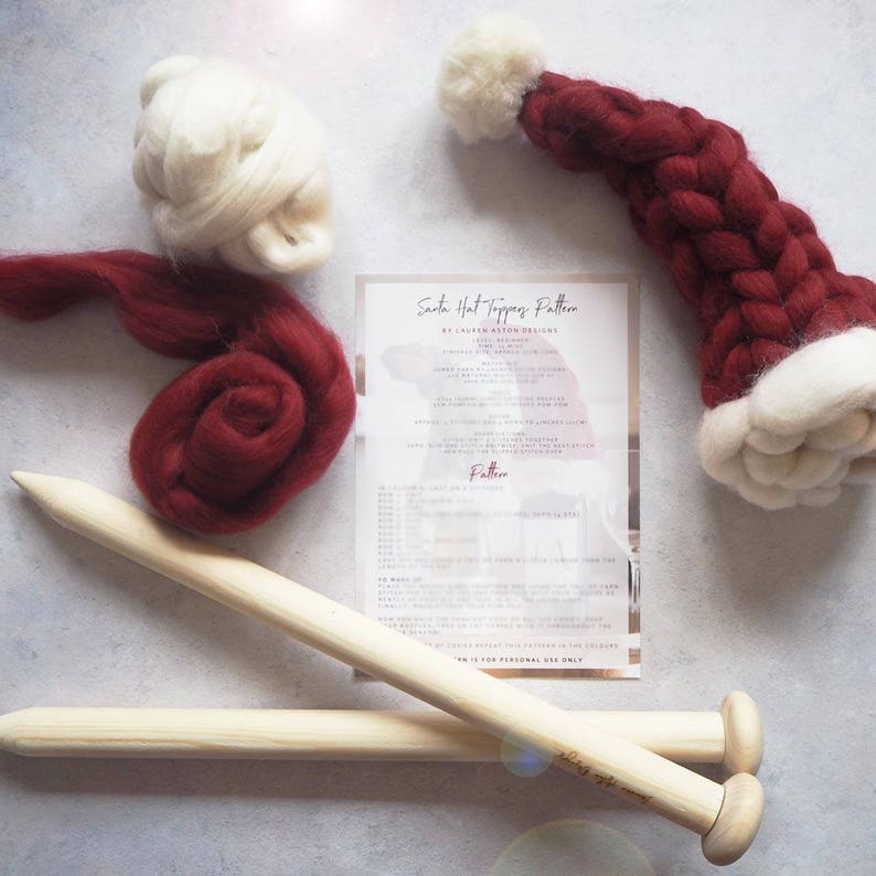 May include: A red and white Santa hat topper knitting kit with instructions, two knitting needles, and red and white wool. The instructions are titled "Santa Hat Toppers Pattern by Laughlin Ashton Designs".