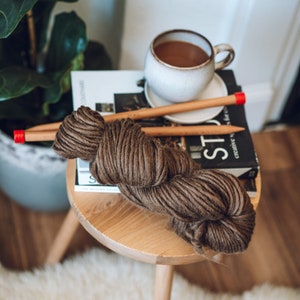 K&ouml;nnte beinhalten: Eine braune Wollkugel mit Holzstricknadeln und einer Tasse Kaffee auf einem Holztisch mit einem Buch mit dem Titel "Creative in Style".