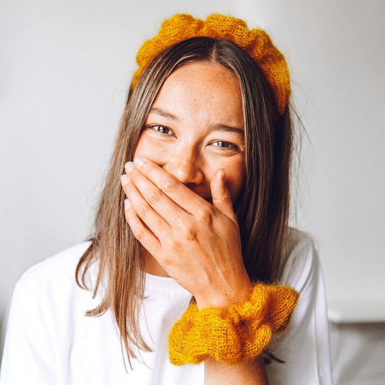 K&ouml;nnte beinhalten: Eine Frau mit langen braunen Haaren lacht und h&auml;lt sich die Hand vor den Mund. Sie tr&auml;gt ein wei&szlig;es Hemd und ein gelbes Strickband.