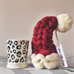 May include: A red and white knitted Santa hat with a white pom pom on top. The hat is sitting on a table next to a white mug with a black leopard print design. The tag on the hat reads "Lauren Ashton Designs".