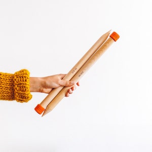 May include: A pair of large wooden knitting needles, each with an orange cap on one end and the measurement "25.00 mm" printed on the side. The needles are held in a hand, with a yellow knitted sleeve visible in the corner.