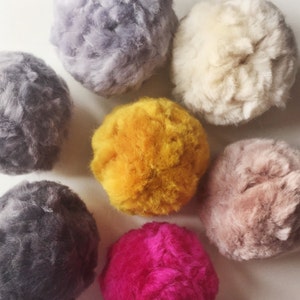 May include: Seven fluffy pom poms in various colors, including gray, white, yellow, pink, and brown. The pom poms are arranged in a circle on a white surface.