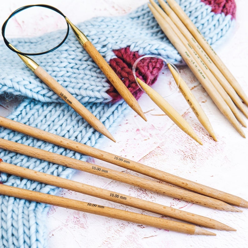 May include: A set of six wooden knitting needles, each measuring 10.00 mm in diameter. The needles are arranged on a white surface with a blue and pink knitted scarf. The needles are labeled with the size in millimeters.