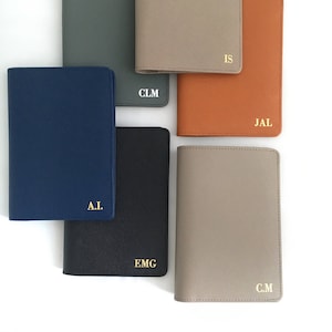 May include: A collection of six rectangular passport holders in navy, dark gray, beige, orange, black, and light gray. Each holder features a textured finish and is personalized with gold or white embossed initials including A.I., CLM, IS, JAL, EMG, and C.M.