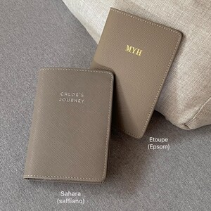 May include: Two taupe passport holders with gold foil lettering. One holder features the text MYH, while the other displays the text CHLOE'S JOURNEY. The holders have distinct textures, with one labeled Etoupe (Epsom) and the other Sahara (saffiano).