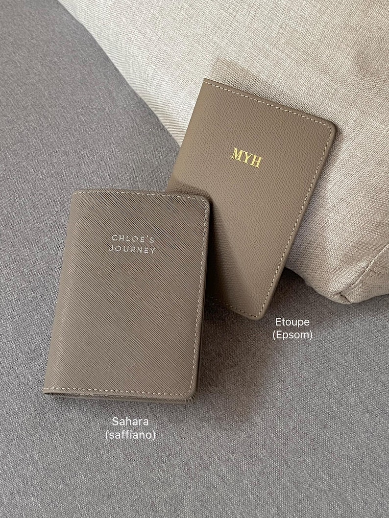 May include: Two taupe passport holders with gold foil lettering. One holder features the text MYH, while the other displays the text CHLOE'S JOURNEY. The holders have distinct textures, with one labeled Etoupe (Epsom) and the other Sahara (saffiano).