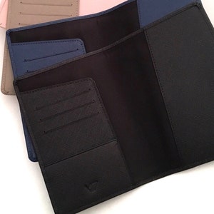 May include: Three open passport holders with a textured crosshatch finish. The front holder is black, the middle is navy blue, and the back is beige. Each holder features interior card slots and a pocket for documents, with a small logo embossed at the bottom.