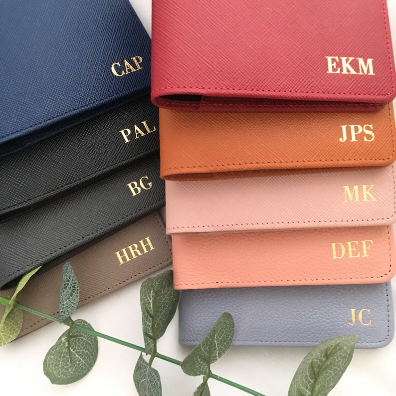 May include: A collection of rectangular wallets in various colors including navy, black, grey, red, orange, pink, peach, and light blue. Each wallet features a textured surface and gold-colored embossed initials: CAP, PAL, BG, HRH, EKM, JPS, MK, DEF, and JC.