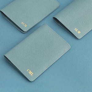 May include: Three light blue passport holders with a textured crosshatch finish. Each rectangular cover features gold foil embossed initials in the bottom corner, specifically the letters ACN, SLW, and CML, arranged in a clean, simple layout.